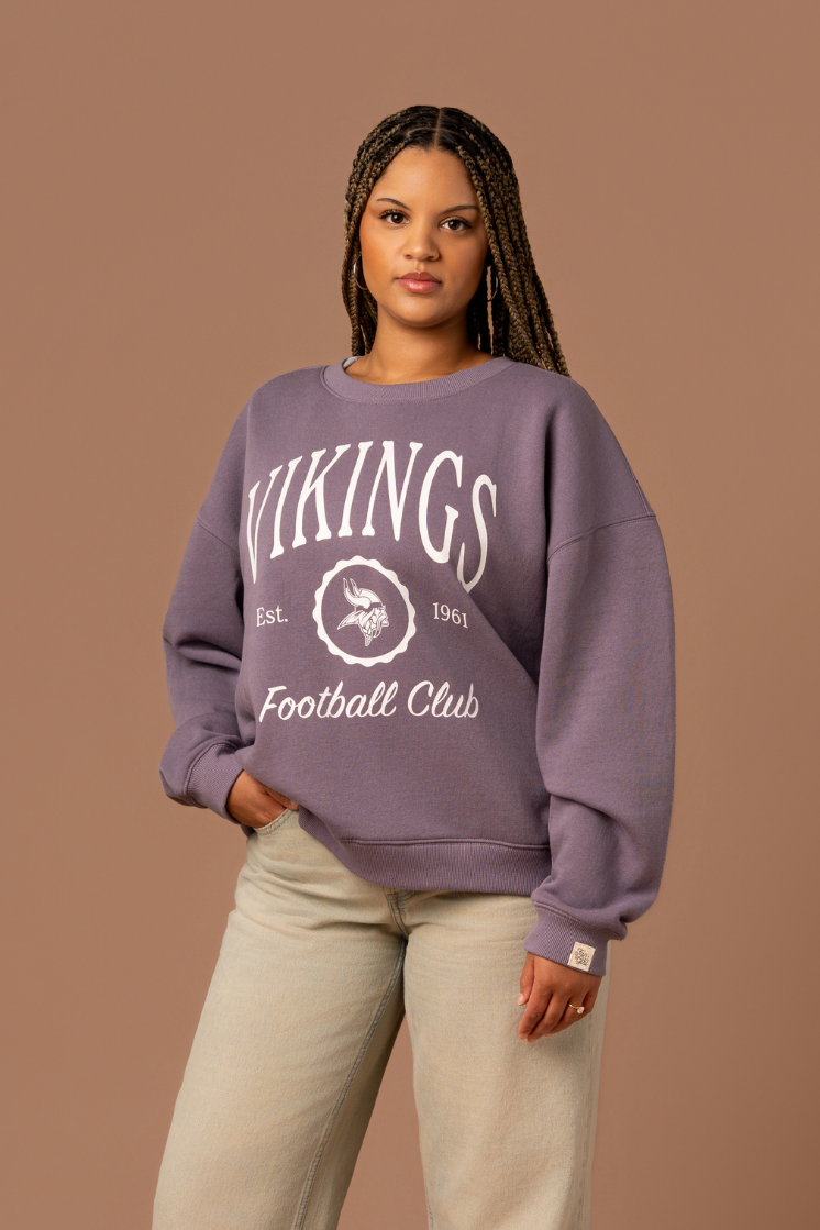 Woman wearing a purple crew neck sweatshirt with white 'Vikings Football Club' and 'EST 1961' text and a white Vikings logo in the center 