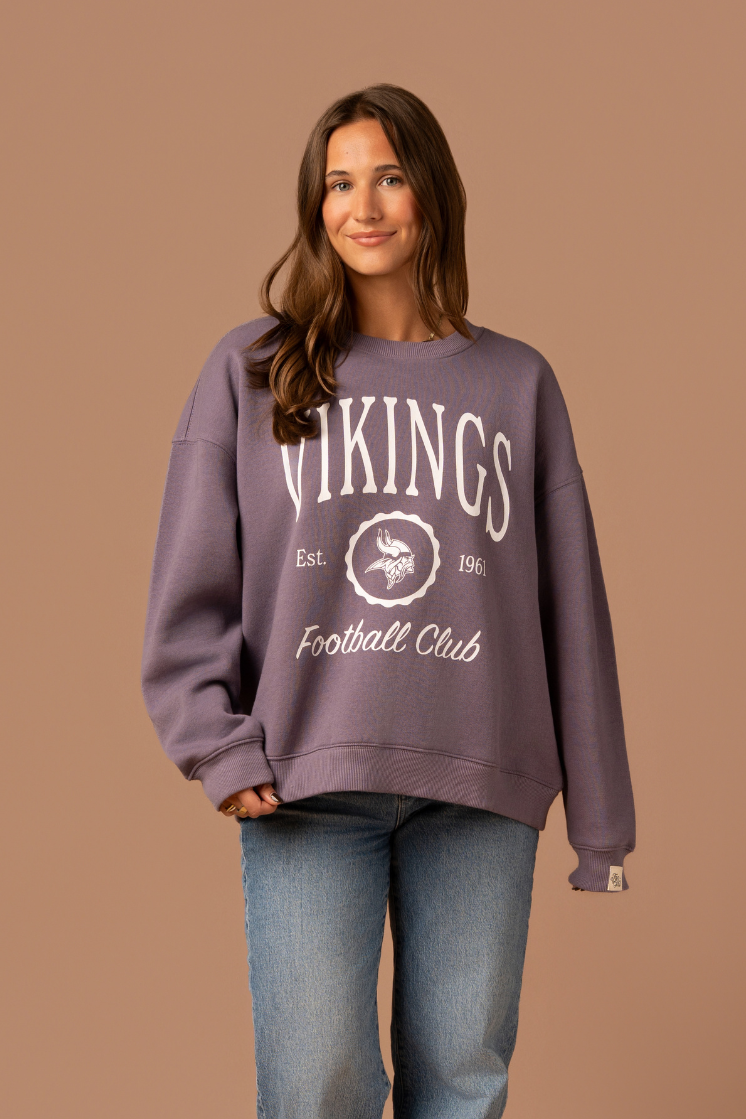 Woman wearing a purple crew neck sweatshirt with white 'Vikings Football Club' and 'EST 1961' text and a white Vikings logo in the center 