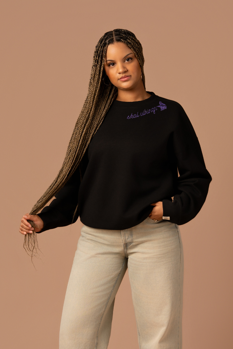 Woman wearing a black crew neck sweatshirt with purple cursive 'Skol Vikings' text and a logo Vikings logo along the neckline 
