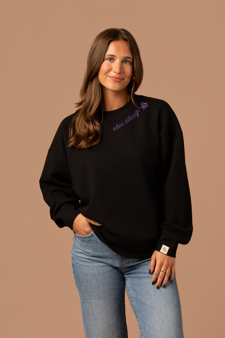 Woman wearing a black crew neck sweatshirt with purple cursive 'Skol Vikings' text and a logo Vikings logo along the neckline 