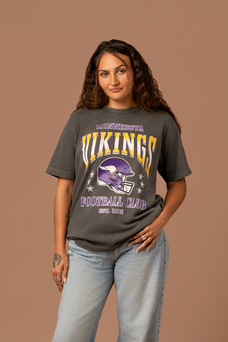 Woman wearing a faded black t-shirt with gold and purple 'Minnesota Vikings Football Club' and 'EST. 1961' text with a purple Vikings helmet graphic in the center 