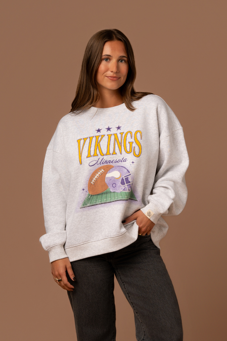 Woman wearing a heather grey crew neck sweatshirt with large yellow 'Vikings' text, purple 'Minnesota' cursive text and a football and helmet graphic under it 