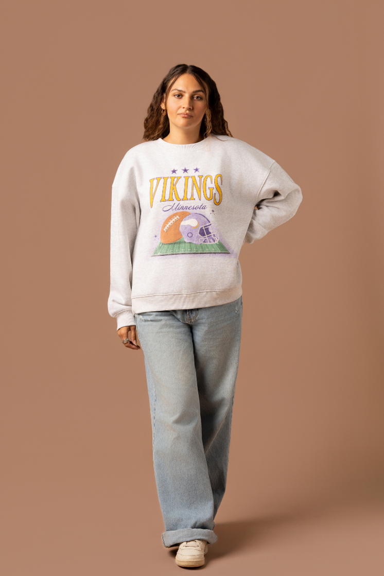 Woman wearing a heather grey crew neck sweatshirt with large yellow 'Vikings' text, purple 'Minnesota' cursive text and a football and helmet graphic under it 