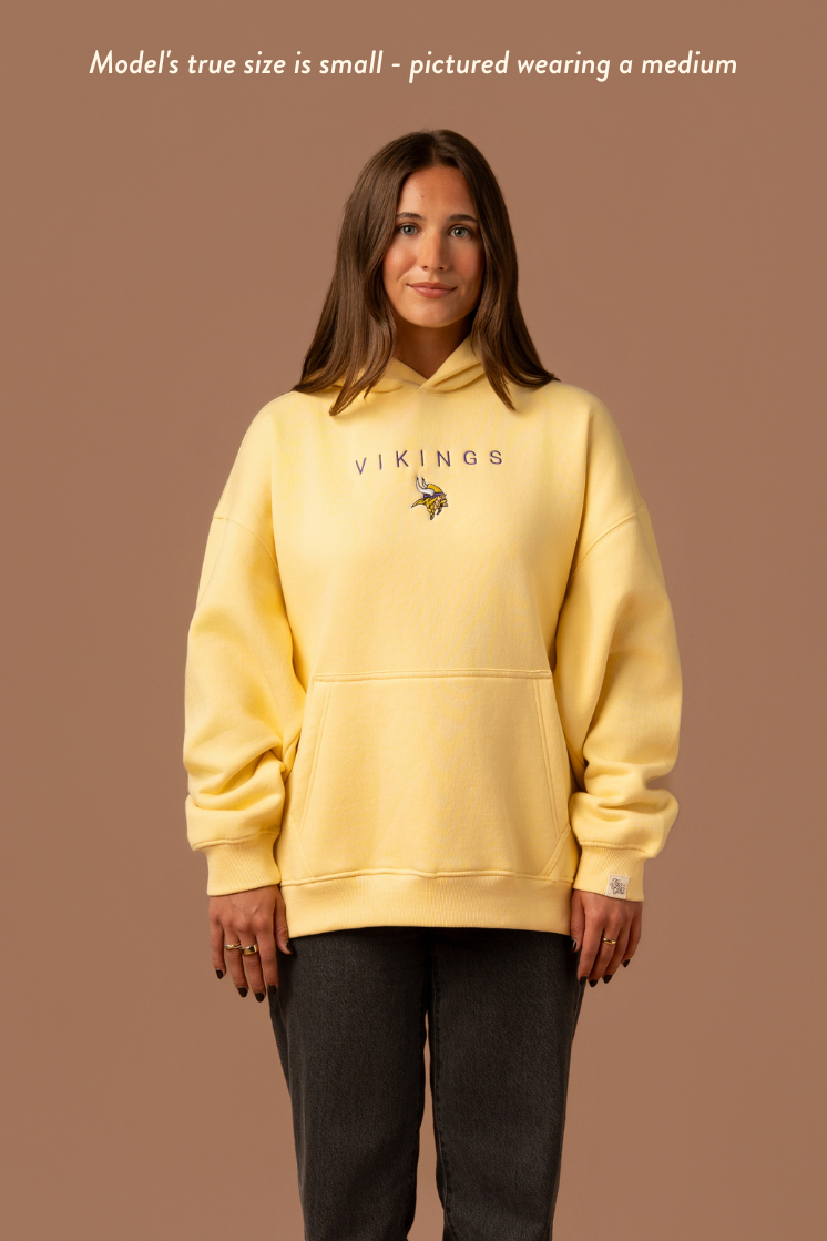 Woman wearing a yellow hoodie with purple 'Vikings' text and a small Vikings logo in the center. Caption reads "model's true size is small- pictured wearing a medium"