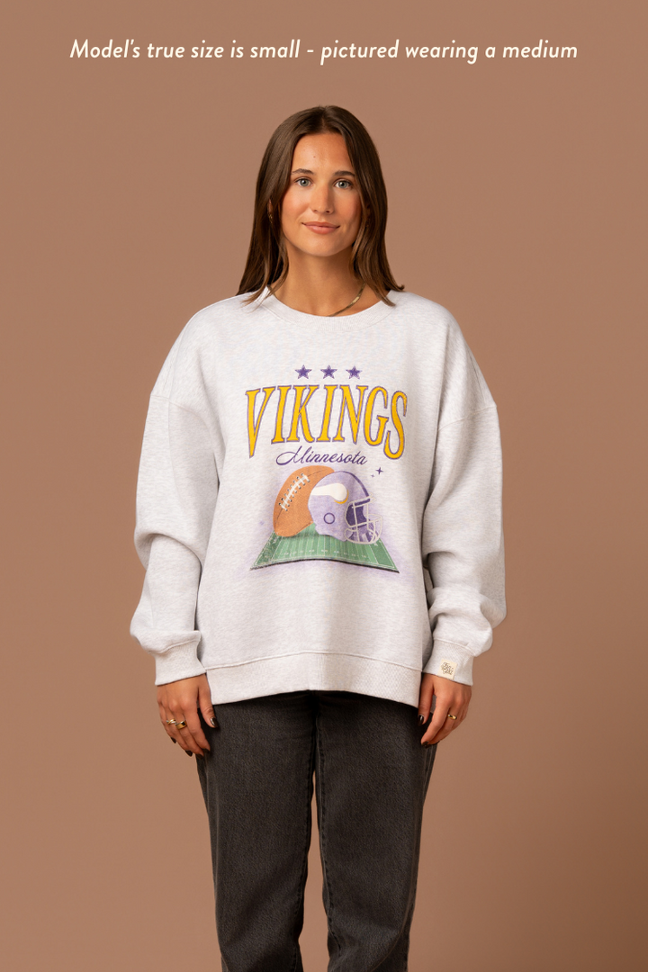 Woman wearing a heather grey crew neck sweatshirt with large yellow 'Vikings' text, purple 'Minnesota' cursive text and a football and helmet graphic under it. Caption reads "Model's true size is small - pictured wearing a medium"