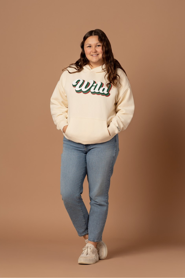 woman standing and farther away of her wild hockey ivory hoodie sweatshirt that is a retro style with her hands in the front pockets