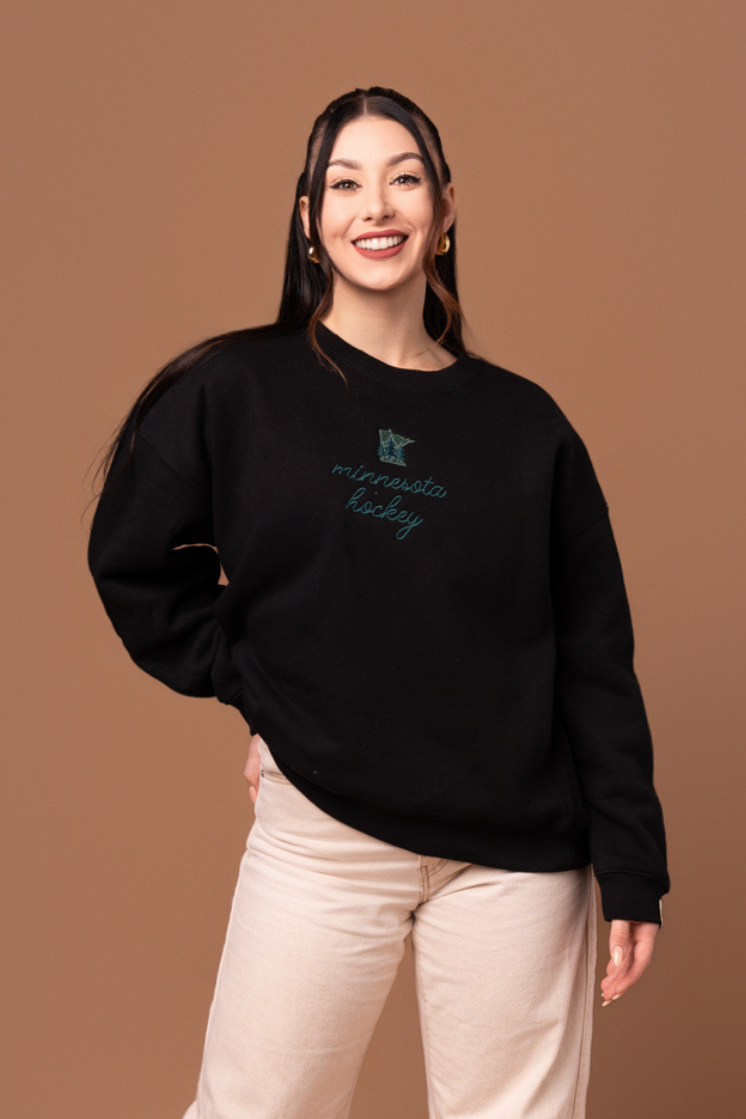 woman in black crew neck sweatshirt with green "minnesota hockey" cursive text and a small green minnesota state graphic with trees on it