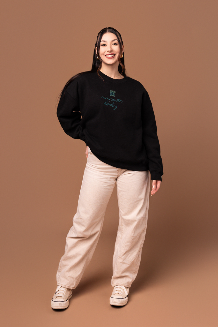 woman in black crew neck sweatshirt with green "minnesota hockey" cursive text and a small green minnesota state graphic with trees on it
