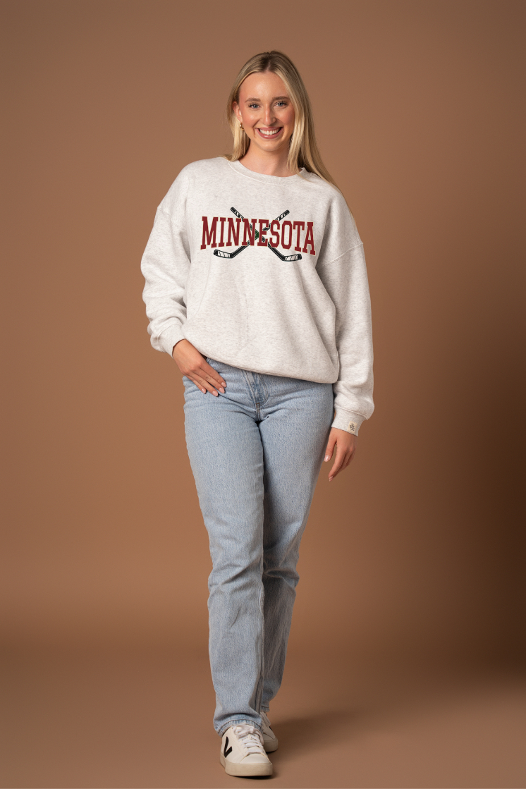 woman in a heather gray crew neck sweatshirt with maroon "minnesota" text and two black hockey sticks