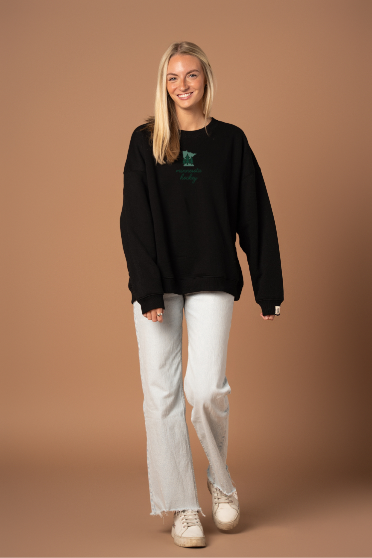 woman in black crew neck sweatshirt with green "minnesota hockey" cursive text and a small green minnesota state graphic with trees on it