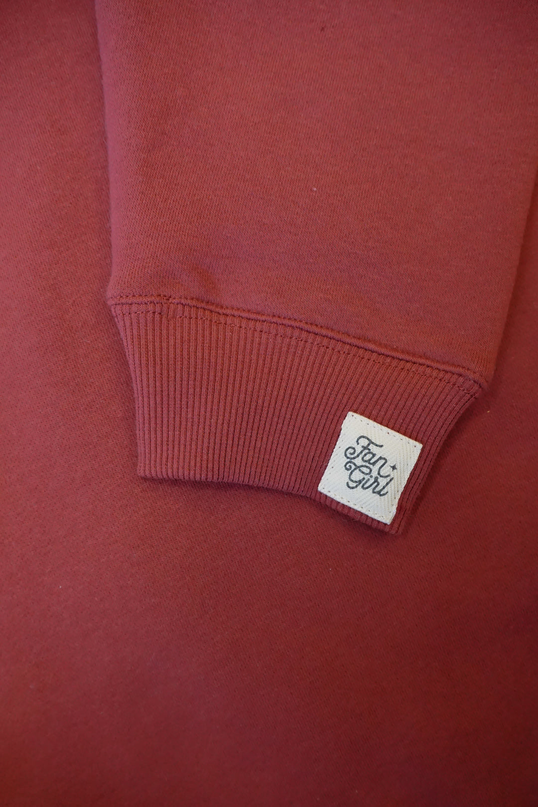 close up image of a maroon sleeve of a crewneck with a Fan Girl logo sewn in