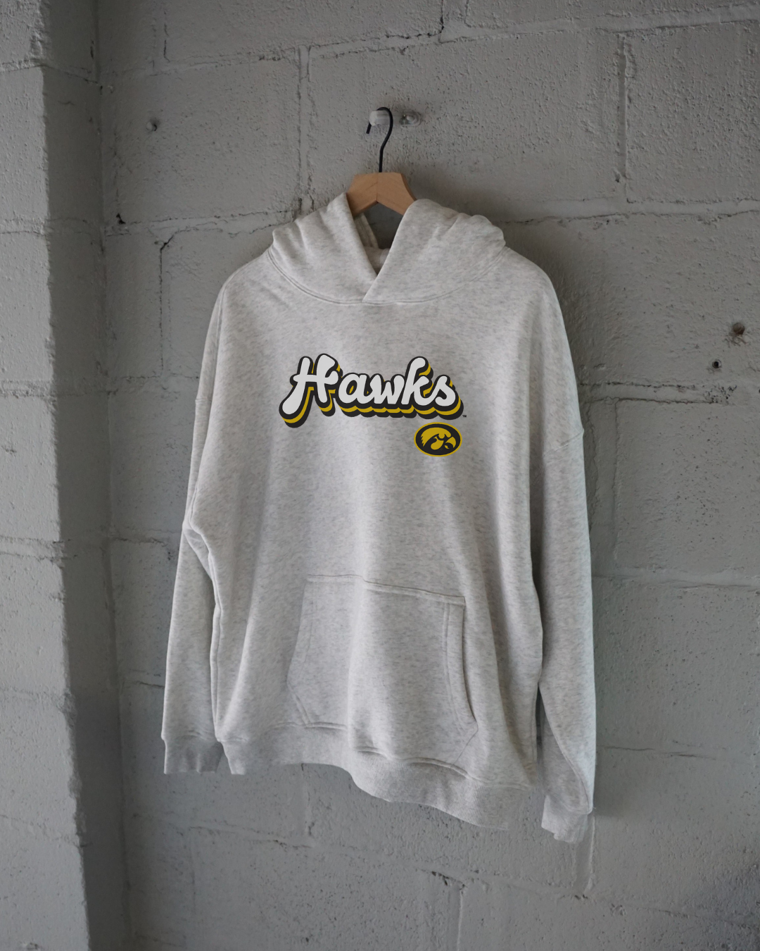 Heather grey hoodie with 'Hawks' text and a Hawkeye logo on a hanger against a wall