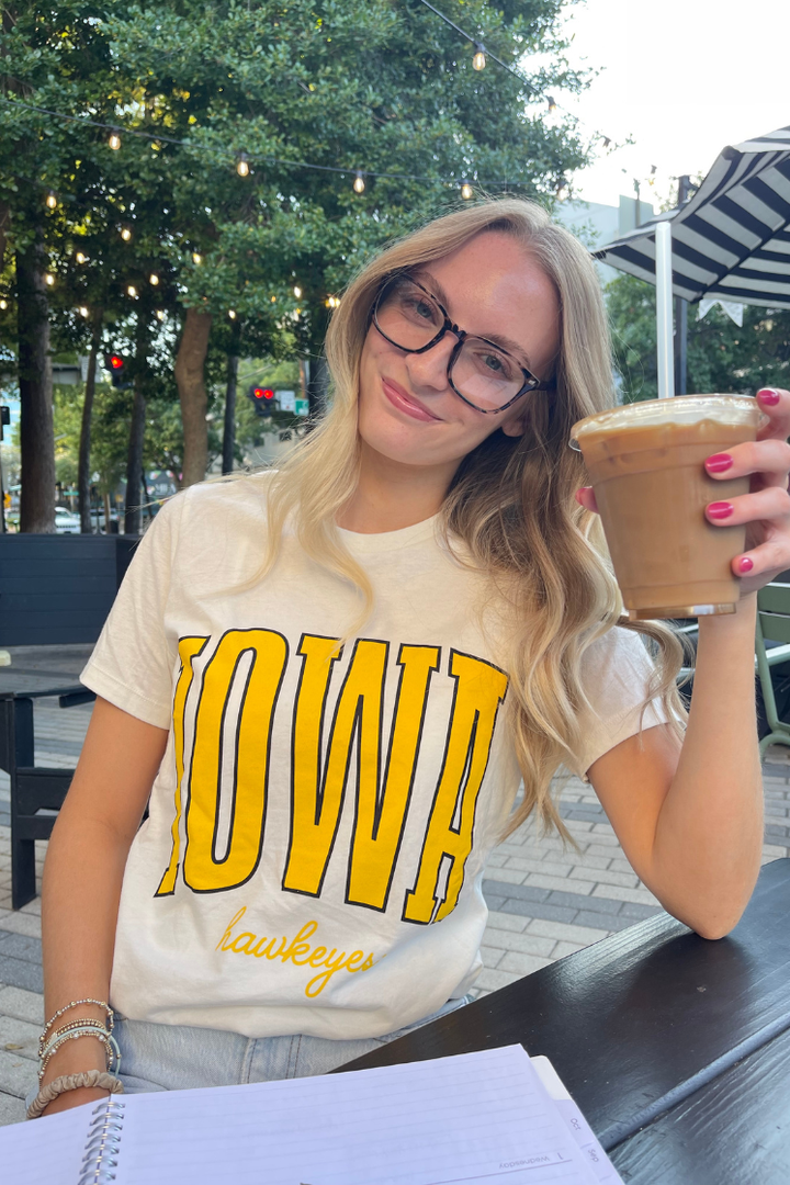 women wearing a white t-shirt with 'Iowa Hawkeyes' text in yellow with a coffee in hand