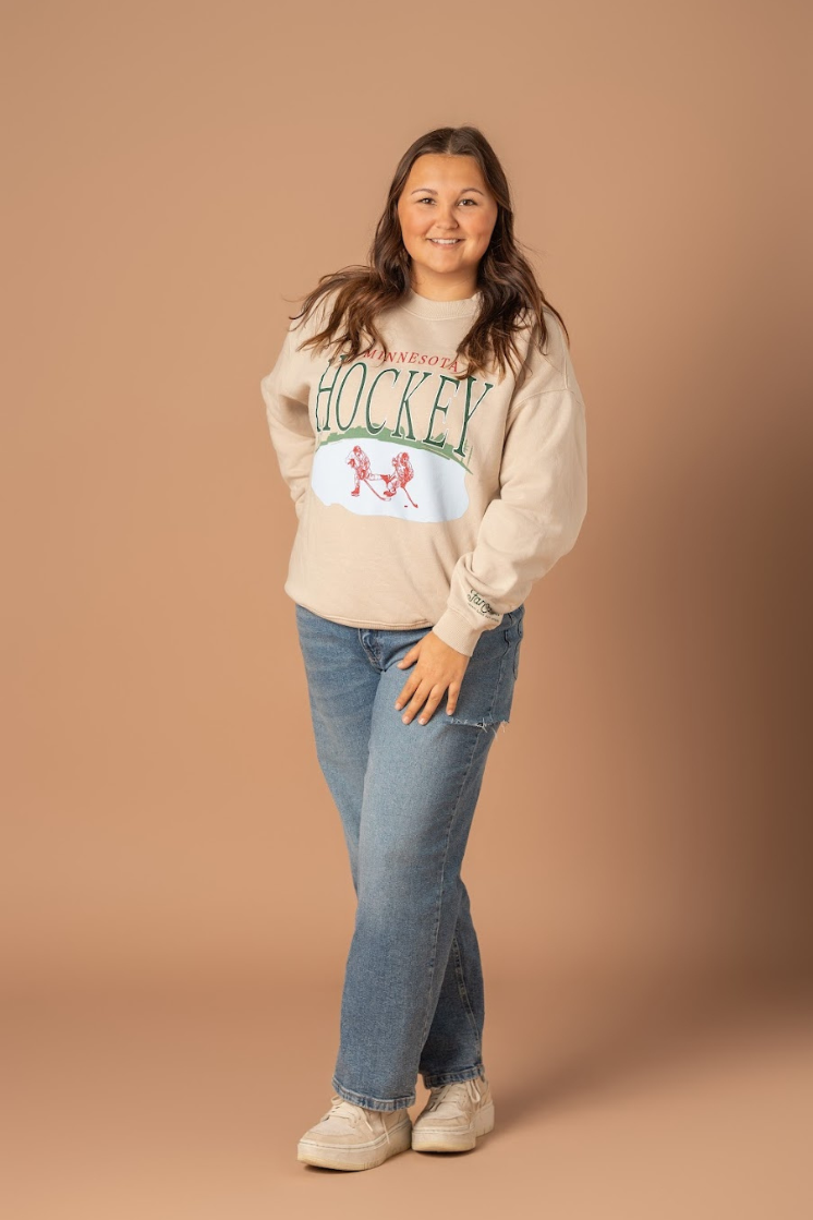 A full body photo of a girl wearing an ivory colored crew neck with a large front design that says Minnesota Hockey with a pond and hockey player graphic. 