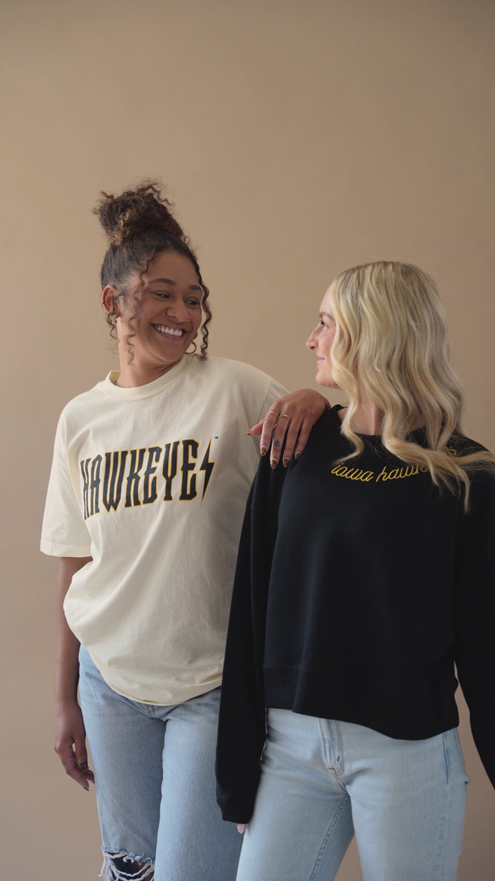 Two girls walking and flaunting their outfit. One wearing an ivory tshirt that has a trademarked Iowa Hawkeyes screenprinted design on it that resembles a trendy band tee. The other in a soft cropped embroidered iowa hawkeyes crew neck.