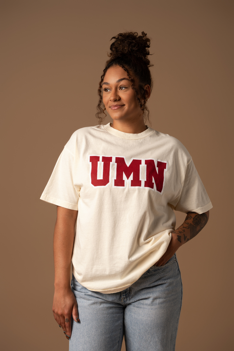 Woman wearing a cream tshirt with UMN screenprinted in block letters in maroon with a white outline