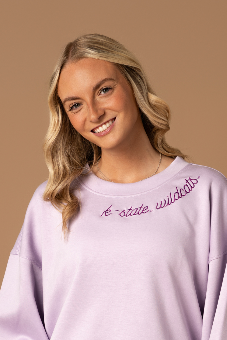 Close up image of a girl wearing a lavender crew neck with an embroidered K-State Wildcats design on the neckline.