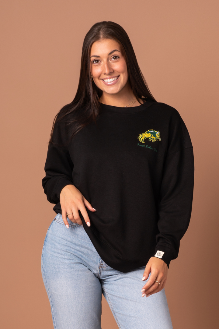 Woman wearing a black full-length crewneck with NDSU logo embroidered in green and gold on the left side 