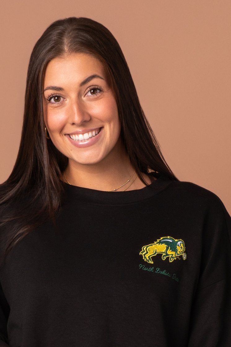 Close up of a woman wearing a black full-length crewneck with NDSU logo embroidered in green and gold on the left side 