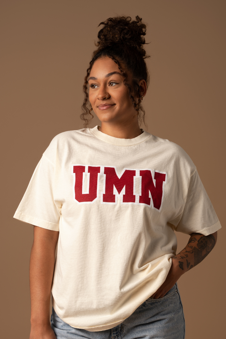 Woman wearing a cream tshirt with UMN screenprinted in block letters in maroon with a white outline