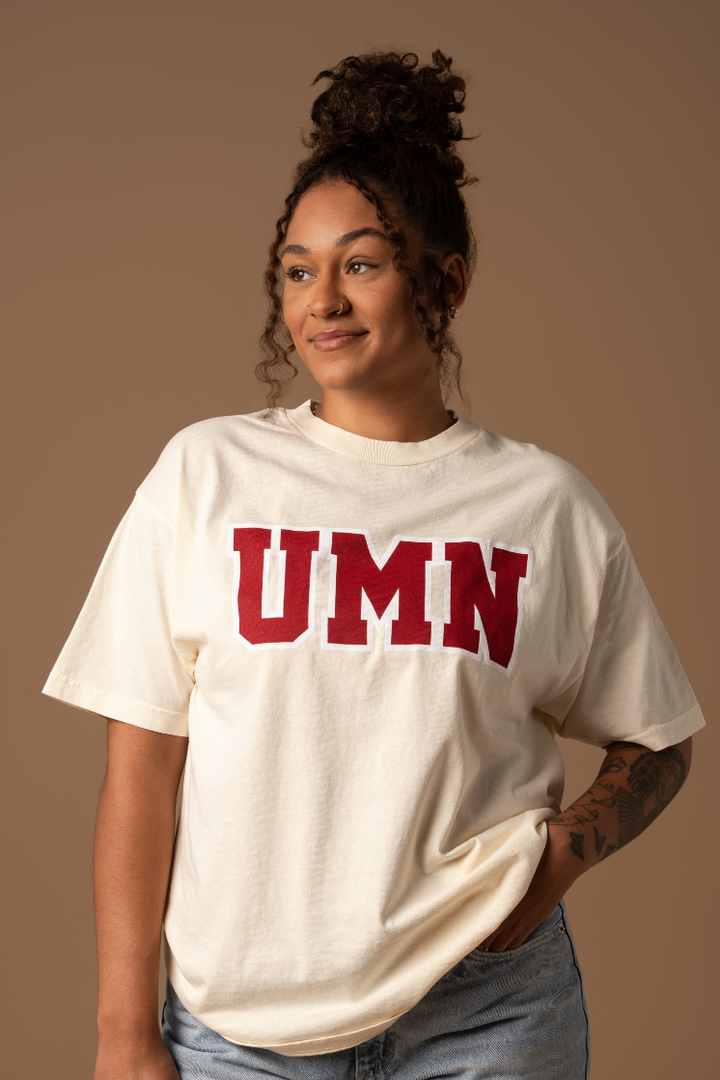 Woman wearing a cream tshirt with UMN screenprinted in block letters in maroon with a white outline