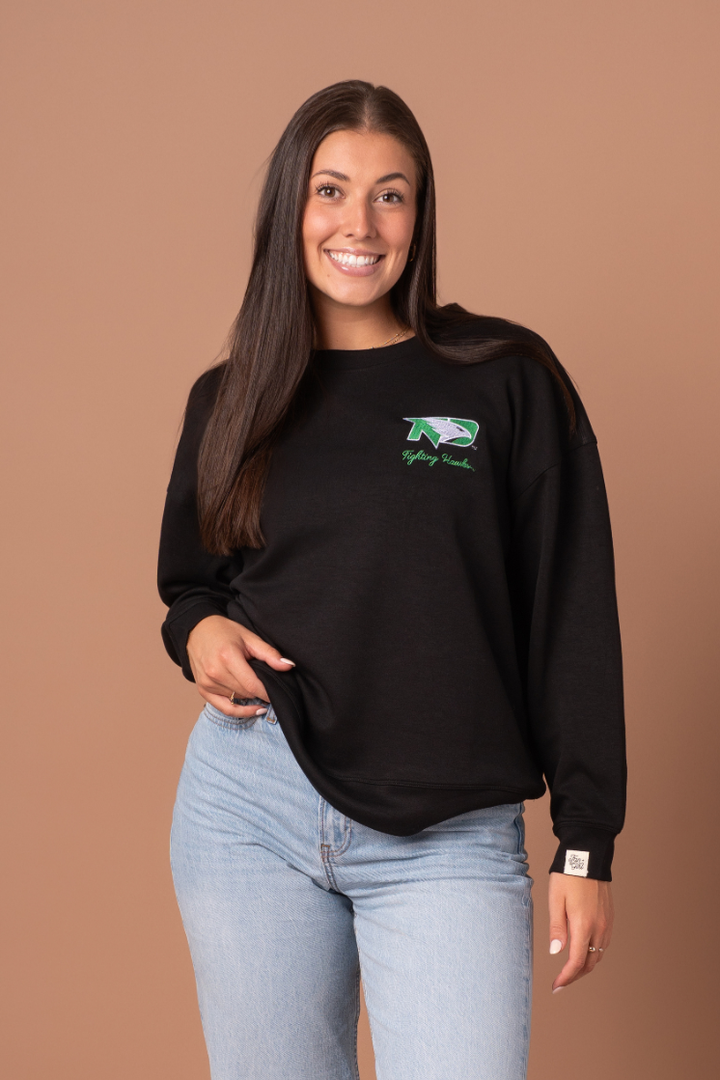 Woman wearing a black crewneck with a UND Fighting Hawk logo embroidered in the left side pocket of the crew