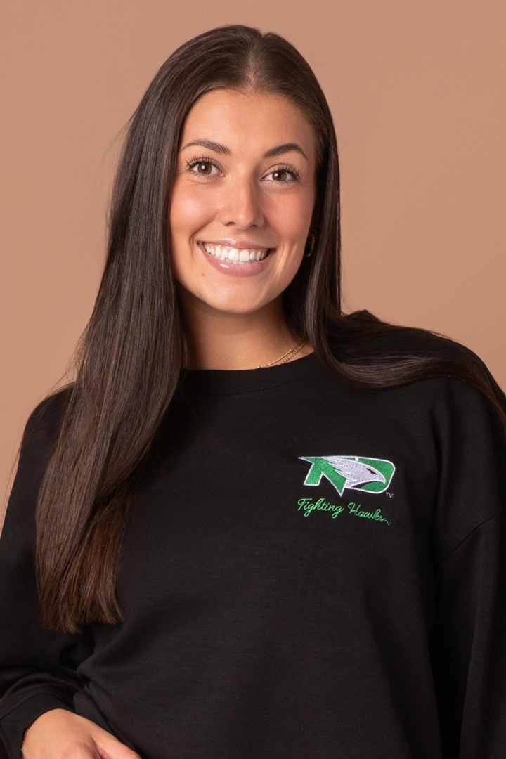 Close up of a woman wearing a black crewneck with a UND Fighting Hawk logo embroidered in the left side pocket of the crew