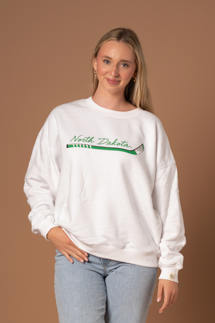 Woman wearing a white crewneck that says North Dakota in kelly green with a green, black and white hockey stick below it