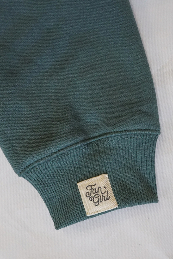 Close-up of a green crew neck sweatshirts sleeve with a branded "Fan Girl" logo