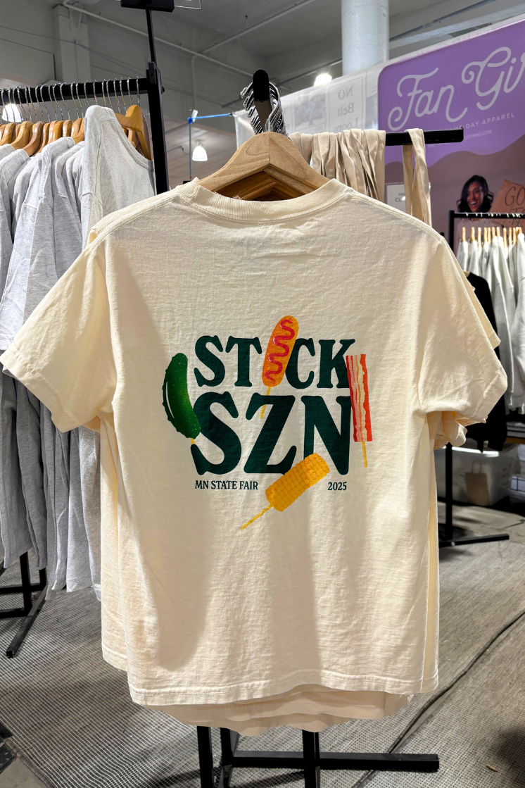 Beige t-shirt with dark green 'stick snz' text and colorful food on-a-stick graphics on a hanger. 