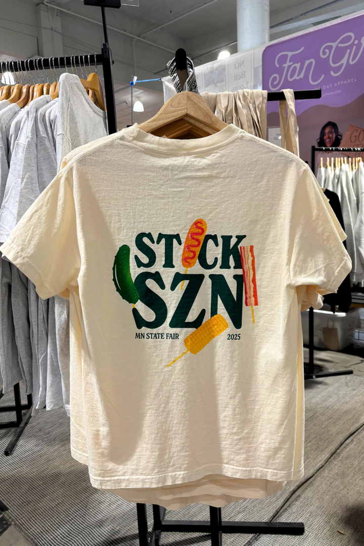 Beige t-shirt with dark green 'stick snz' text and colorful food on-a-stick graphics on a hanger. 