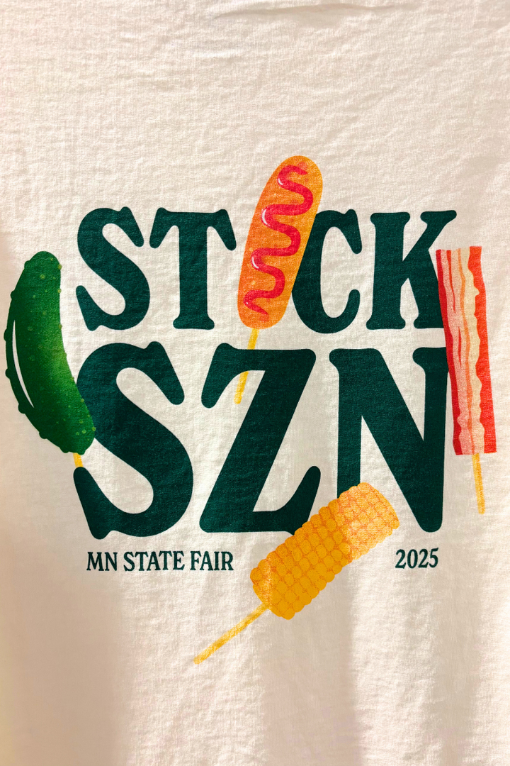 Beige t-shirt with dark green 'stick szn' text and colorful food on-a-stick graphics. 