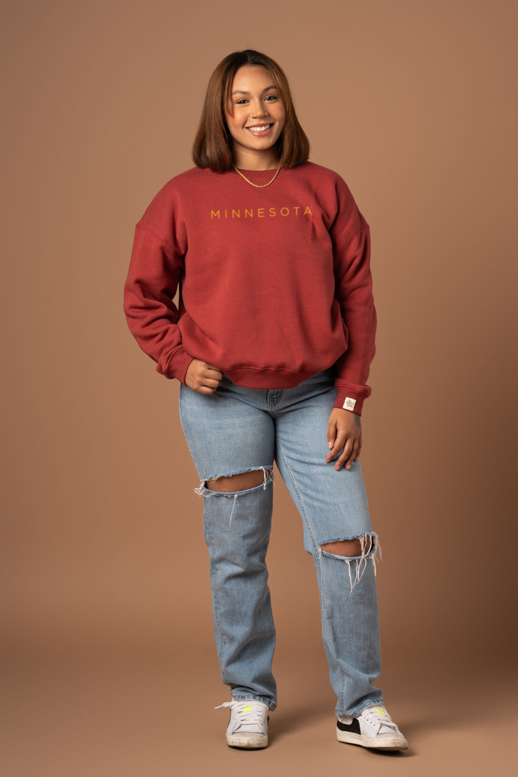 Woman wearing a maroon 'MINNESOTA' crew neck sweatshirt against a brown background