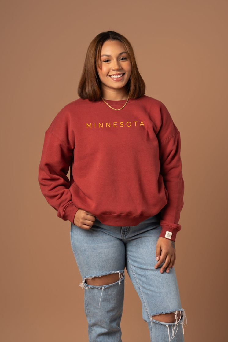 Woman wearing a maroon 'MINNESOTA' crew neck sweatshirt against a brown background